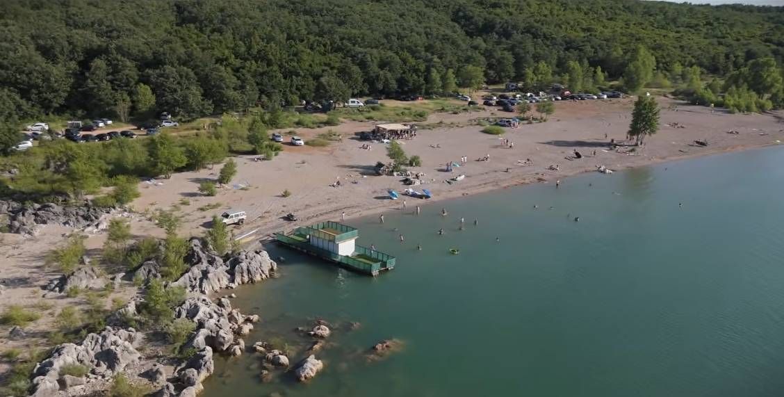 Pogled iz zraka na plažu Buškog jezera s kupačima, stjenovitom obalom i šumom u pozadini.