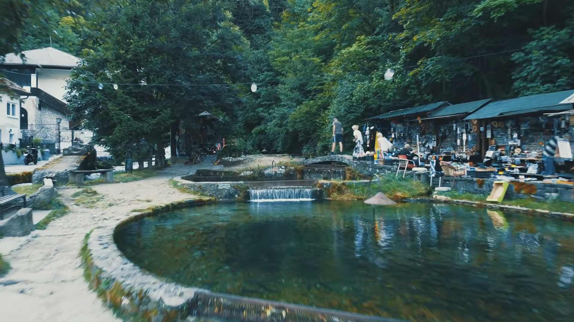 Plava voda u Travniku s malim slapom, kamenim šetnicama i ugostiteljskim objektima uz vodu.